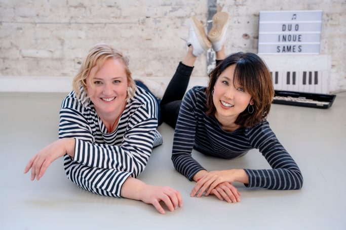 Mari Inoue und Katharina Sames liegen auf dem Boden. Im Hintergrund sieht man eine Klaviertastatur und eine Querflöte